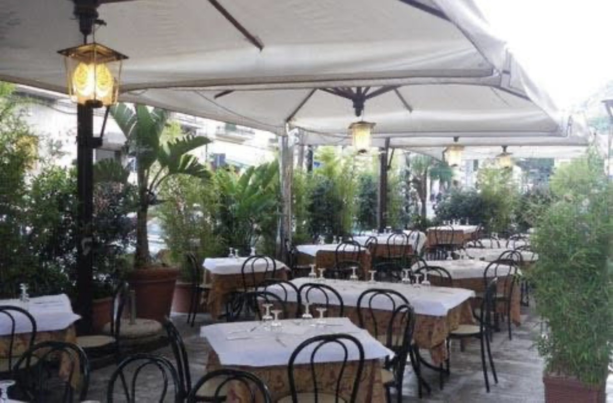 Tavoli all'aperto sotto ombrelloni bianchi, tra piante e luci a lanterna